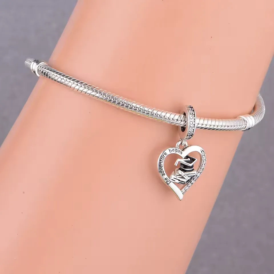 Celebrate new beginnings with this stunning Graduation Dangle Charm! Designed in&nbsp;sterling silver, this elegant charm features an open-heart frame engraved with "The Adventure Begins", surrounding a detailed graduation cap, diploma, and books. The charm is beautifully accented with sparkling Cubic Zirconia stones and black enamel for added contrast and sophistication. Perfect for graduates stepping into their next chapter.